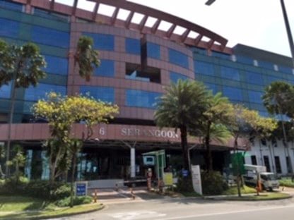 A five story building with a red brick exterior and horizontal glass windows, showing the front of the building with the main entrance. There are seven palm trees visible and the words 6 Serangoon on the building.